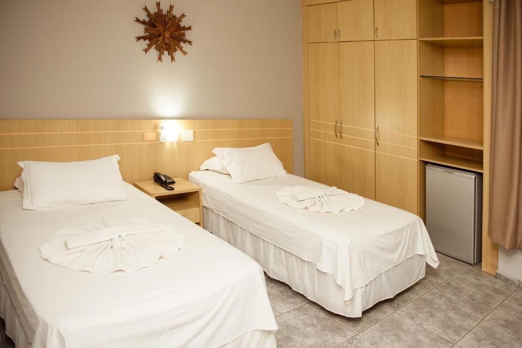 A hotel room with two single beds, a wooden wardrobe, a desk with a telephone, and a small refrigerator.