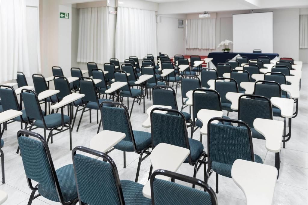 Uma sala de conferência com fileiras de cadeiras e mesas, uma tela de projetor e um pódio.