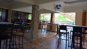 A spacious indoor area with multiple tables and chairs, a fan on the ceiling, and a view of greenery outside. - Diplomat Diani Beach Resort (Diani Beach)