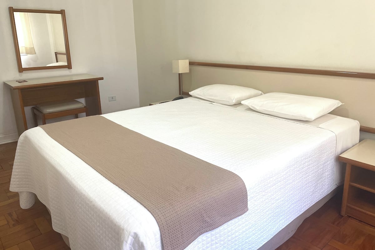 Een opgemaakt bed met een wit-beige dekbed, een houten nachtkastje, een bureau met stoel, en een spiegel aan de muur.