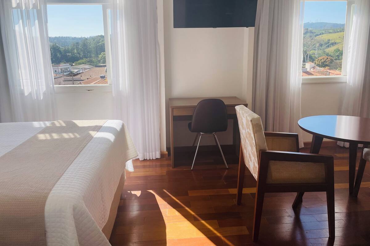 Een hotelkamer met een bed, een bureau, een stoel, een ronde tafel en grote ramen met uitzicht op het landschap.