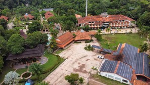 Eine Vogelperspektive zeigt einen Ferienort mit mehreren Gebäuden, darunter ein großes Haupthaus mit rotem Dach, ein kleineres Gebäude mit Reetdach und ein Bauwerk mit Solaranlage.