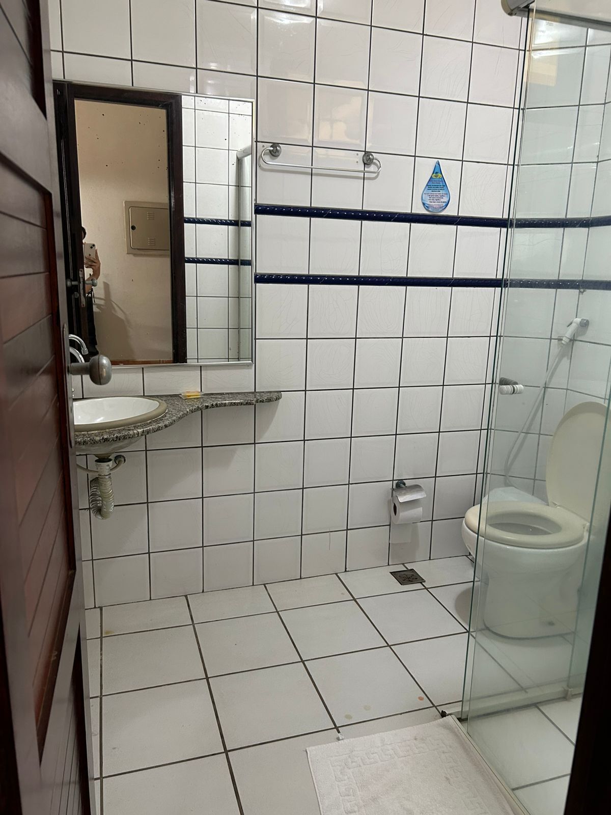 Casa de banho com azulejos brancos, uma pia, sanita e área de duche com cabine de vidro.