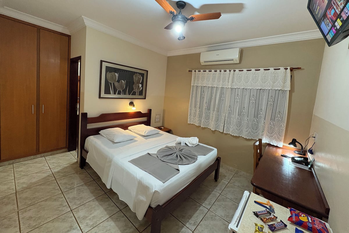 Une chambre bien rangée, avec un lit, un bureau, un ventilateur de plafond, un climatiseur fixé au mur et un tableau encadré au-dessus du lit.
