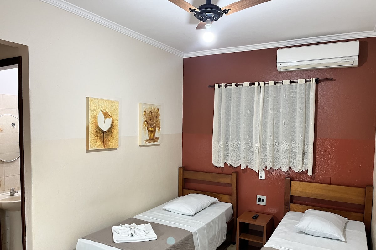 Une chambre d’hôtel avec deux lits simples, une tête de lit en bois, une petite table de chevet, un appareil de climatisation et deux œuvres d’art encadrées accrochées au mur.
