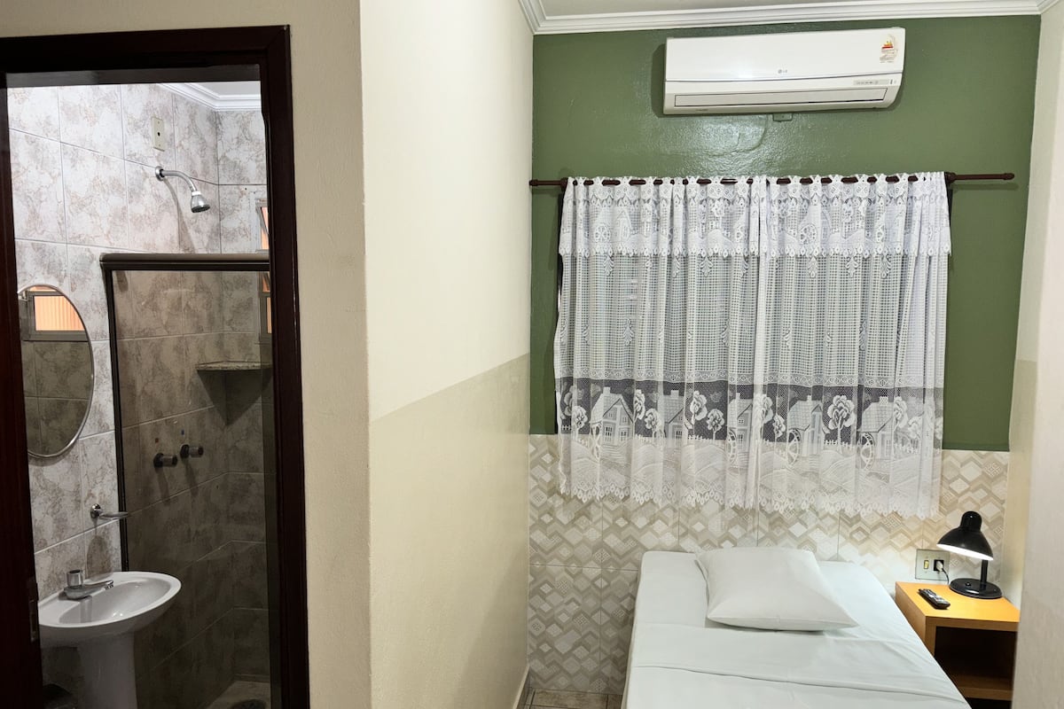 Une salle de bain avec une douche, un lavabo et des toilettes. On y trouve un lit avec un oreiller, une table de chevet avec une lampe et une télécommande.