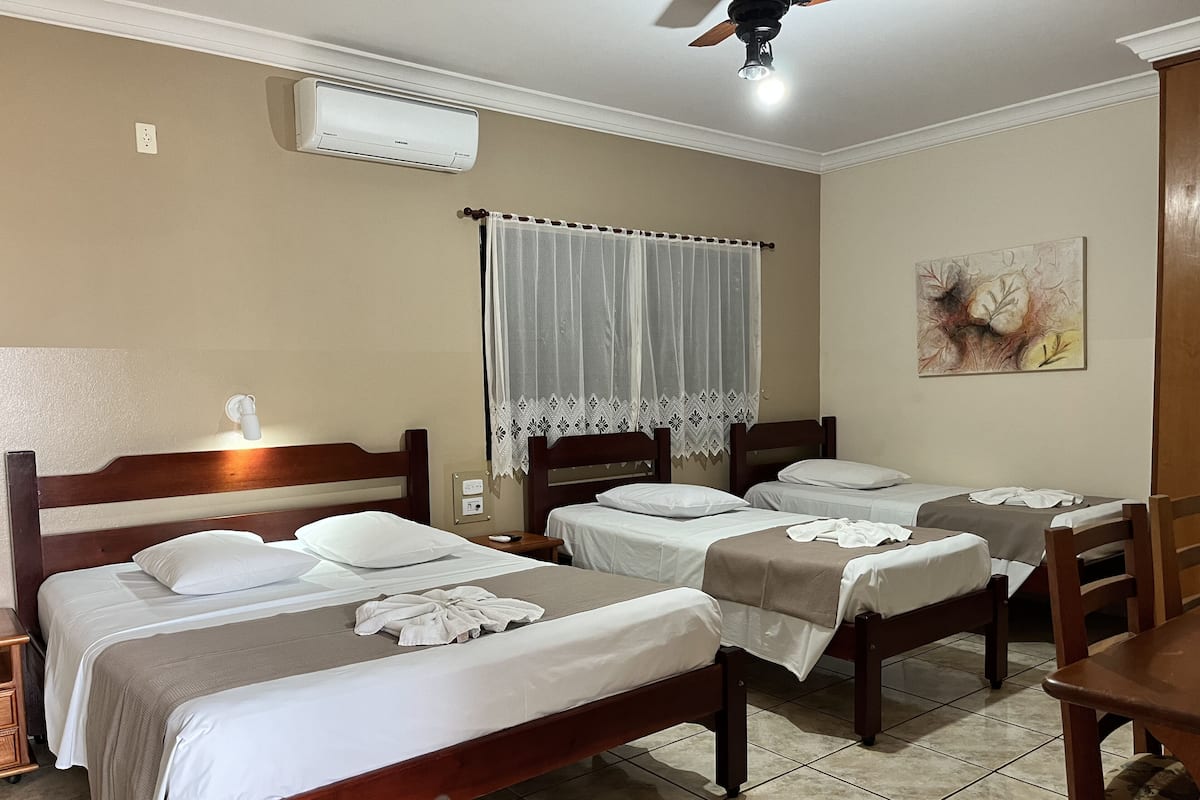 Une chambre d’hôtel avec deux lits, un ventilateur de plafond, un appareil de climatisation, une fenêtre avec un rideau en dentelle et un tableau au mur.