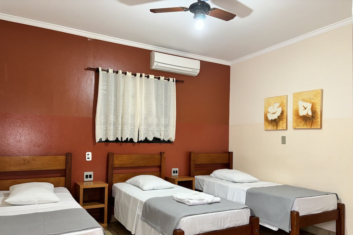 Une chambre d’hôtel avec quatre lits simples, un ventilateur de plafond, un appareil de climatisation, une fenêtre avec des rideaux et deux tableaux représentant des fleurs, accrochés au mur.