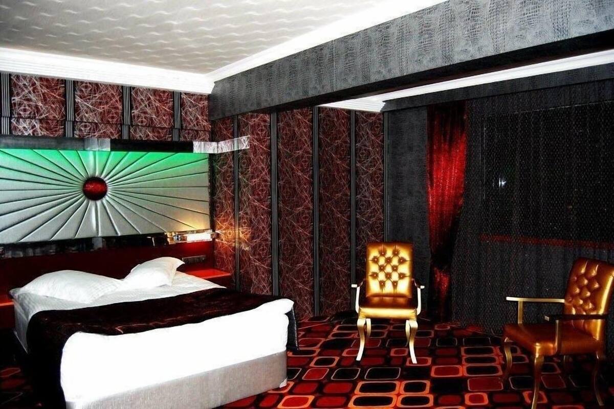 A modern hotel room with a large bed, two armchairs, a patterned carpet, and a unique wall design.