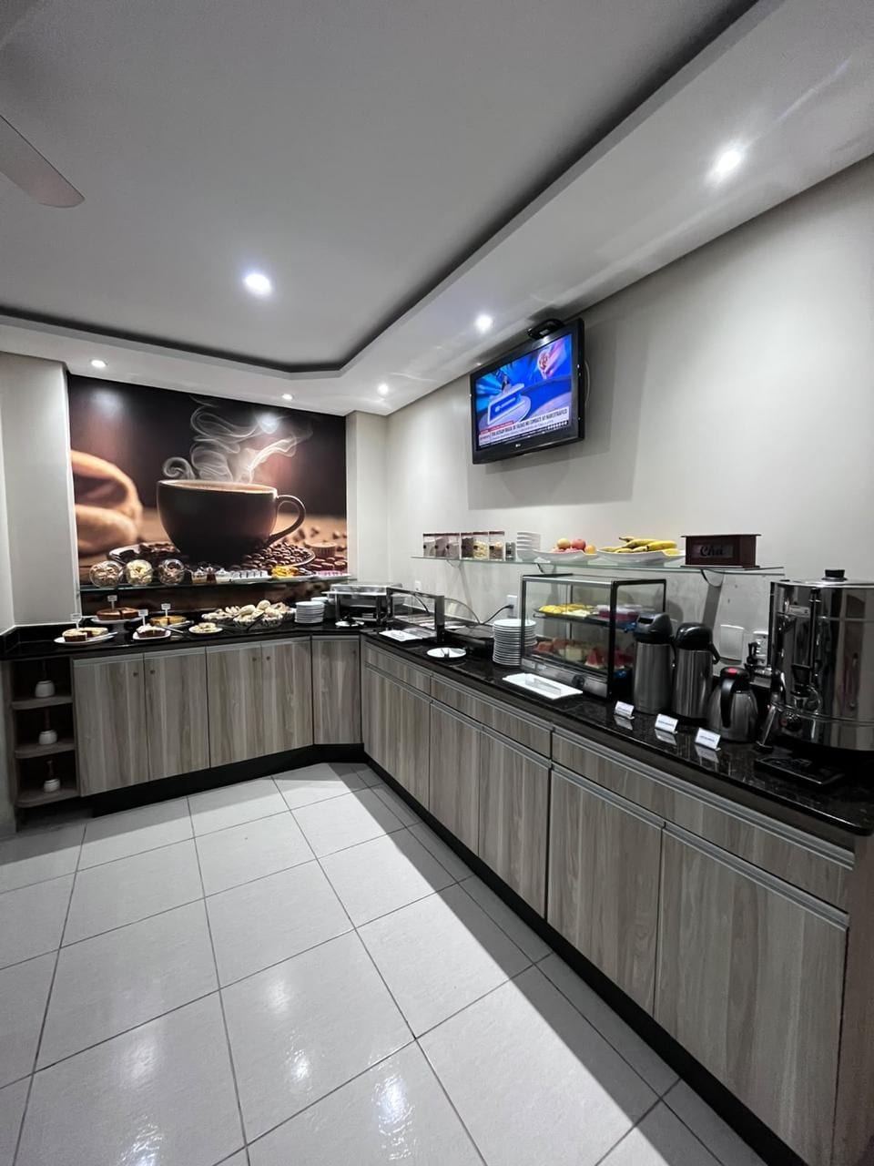A modern kitchen area with a coffee station, a flat-screen TV mounted on the wall, and a tiled floor.