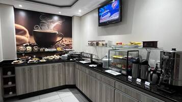 A modern kitchen area with a coffee station, a flat-screen TV mounted on the wall, and a tiled floor.