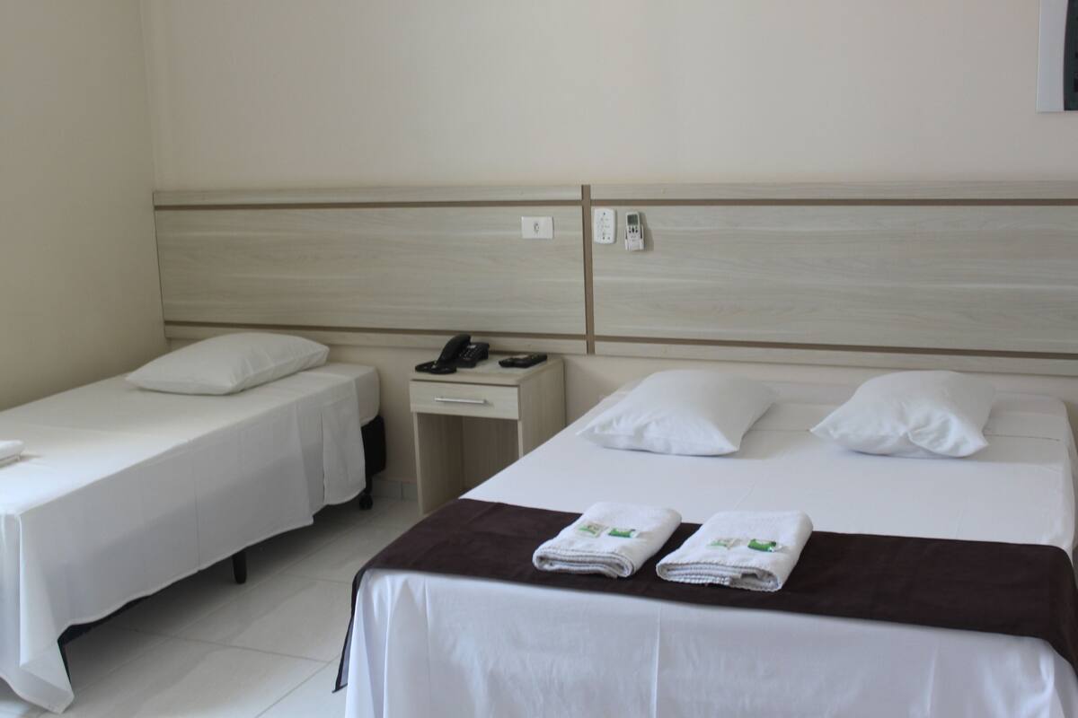 A hotel room with two single beds, a larger bed, a bedside table with a phone, and a wall-mounted control panel.