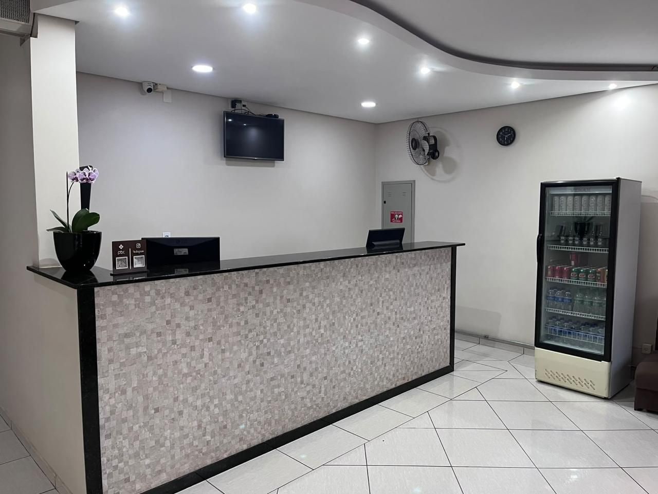 Uma área de recepção com piso ladrilhado, balcão de atendimento, televisão, relógio, ventilador e máquina de vendas.