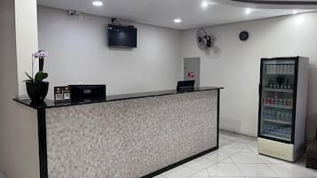 Uma área de recepção com piso ladrilhado, balcão de atendimento, televisão, relógio, ventilador e máquina de vendas.