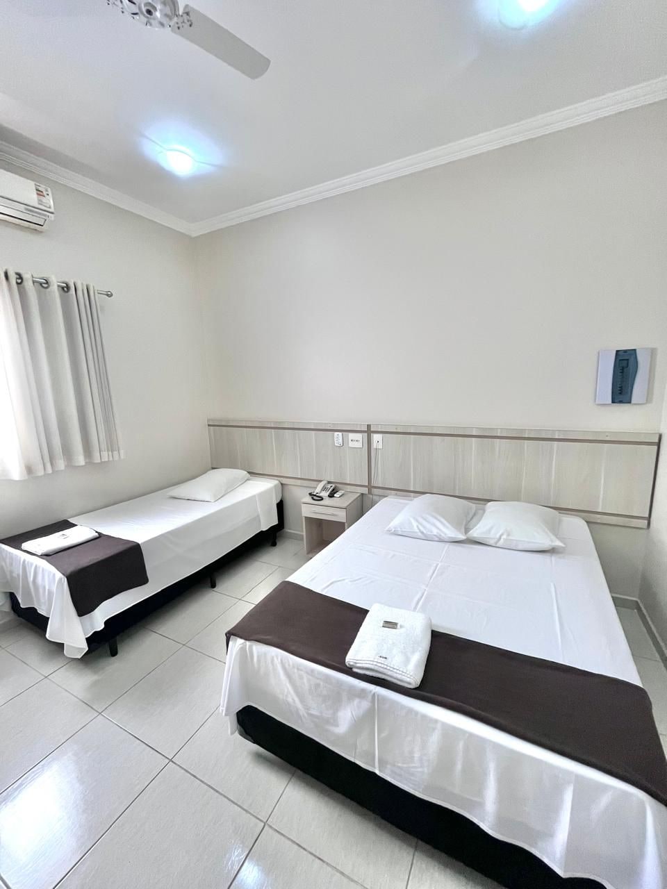 Habitación de hotel con dos camas individuales, una mesita de noche, un ventilador de techo y una ventana con cortinas.