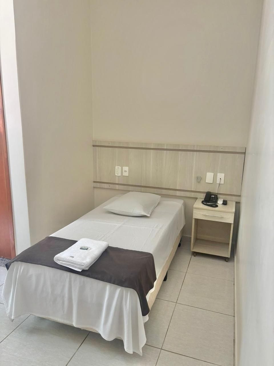 A hotel room with a single bed, a bedside table with a phone, and a wall-mounted light.