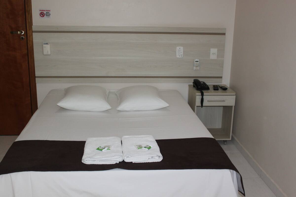 A hotel room with a bed, two pillows, a bedside table with a phone and remote controls, and two folded towels on the bed.