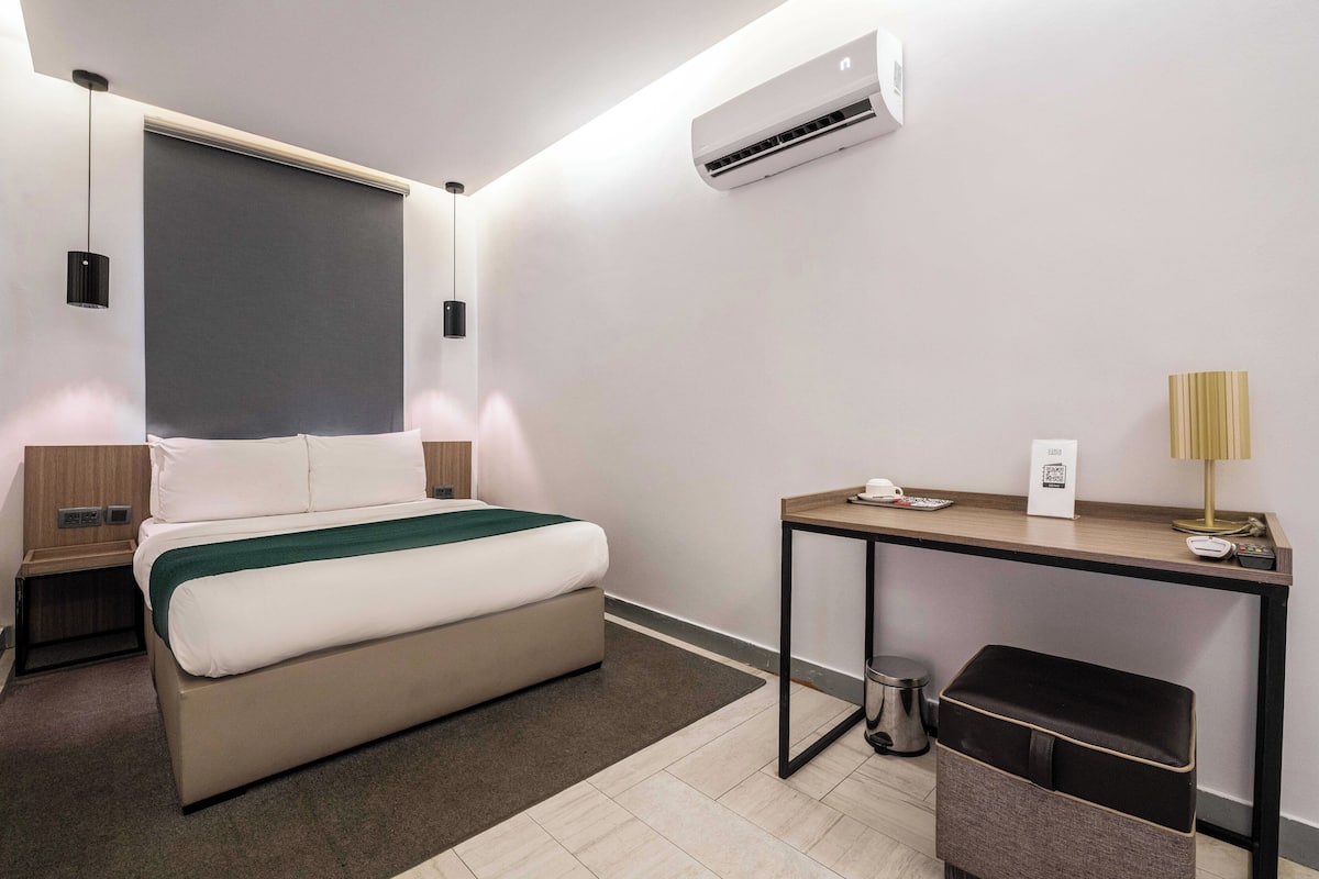 A modern hotel room with a bed, bedside table, desk, and an air conditioning unit.