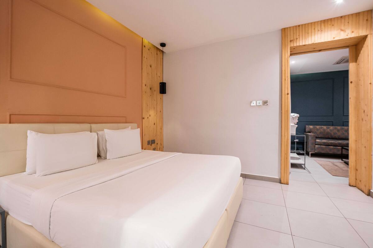 A bedroom with a large bed, wooden paneling on the walls, and a view into another room with a sofa.