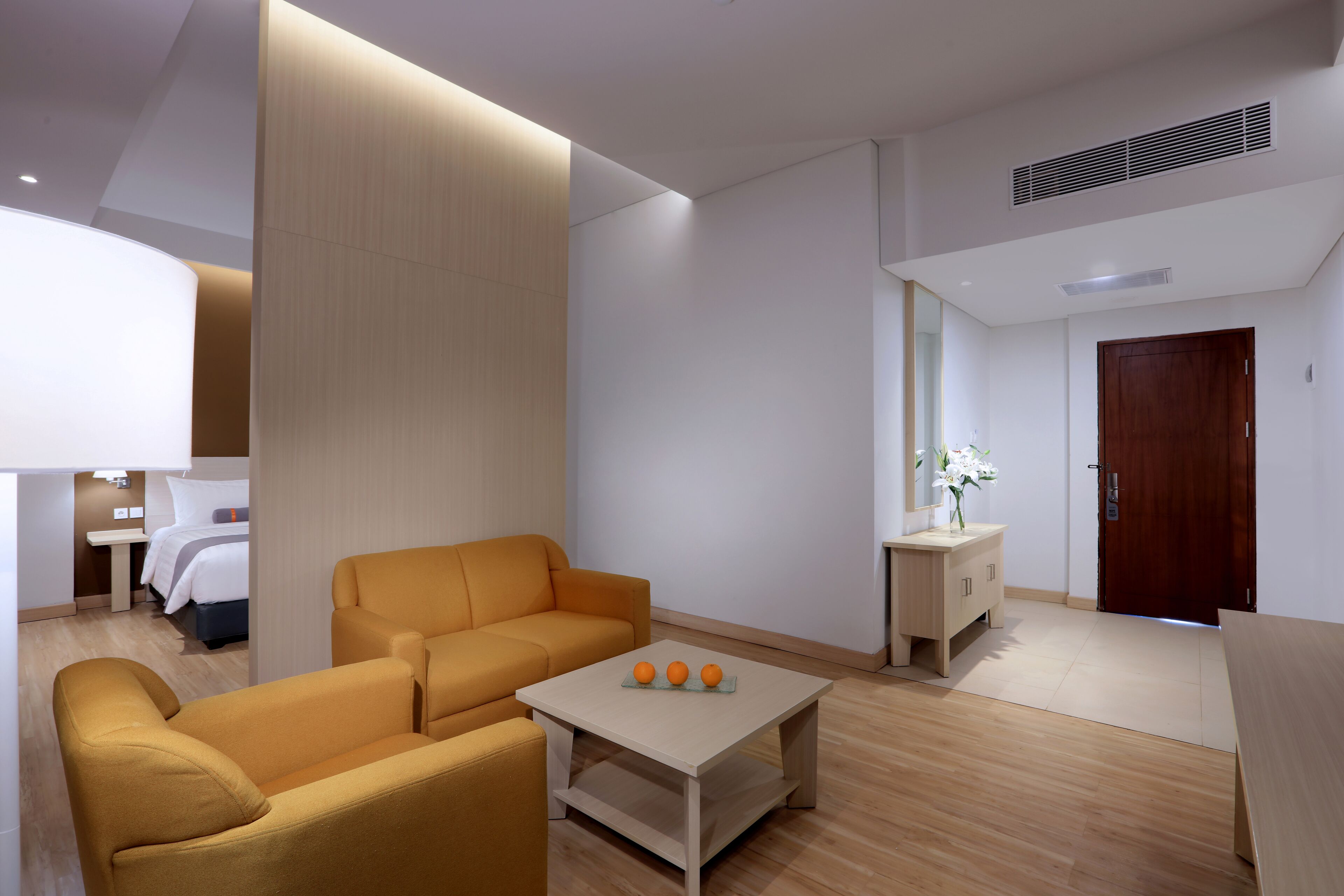 A modern hotel room with a sofa, a small table with oranges, a bedside table, a mirror, and a wooden door.