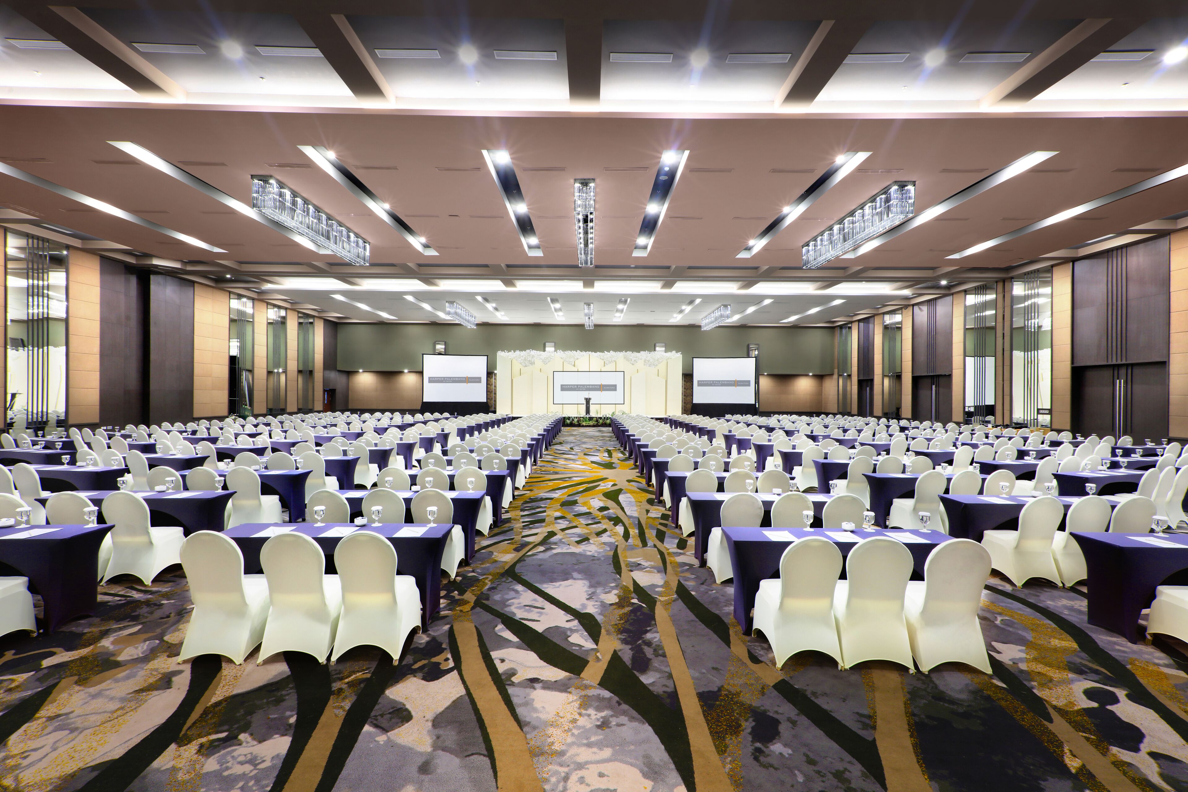 Un salón de conferencias con filas de mesas y sillas, suelo alfombrado y un escenario al frente.