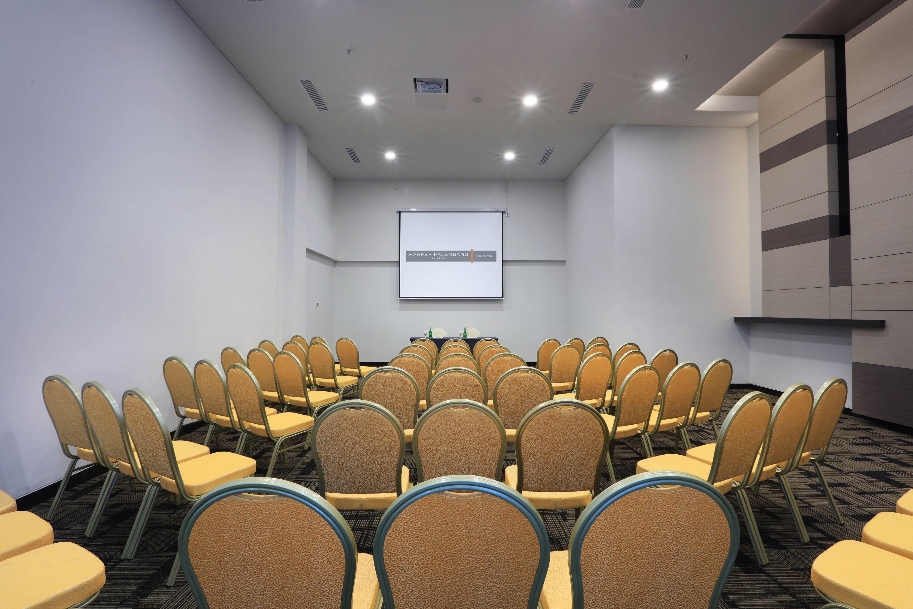 Un salón de conferencias con filas de sillas orientadas hacia una pantalla de proyección, una mesa con botellas de agua y suelo alfombrado.