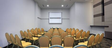 Un salón de conferencias con filas de sillas orientadas hacia una pantalla de proyección, una mesa con botellas de agua y suelo alfombrado.