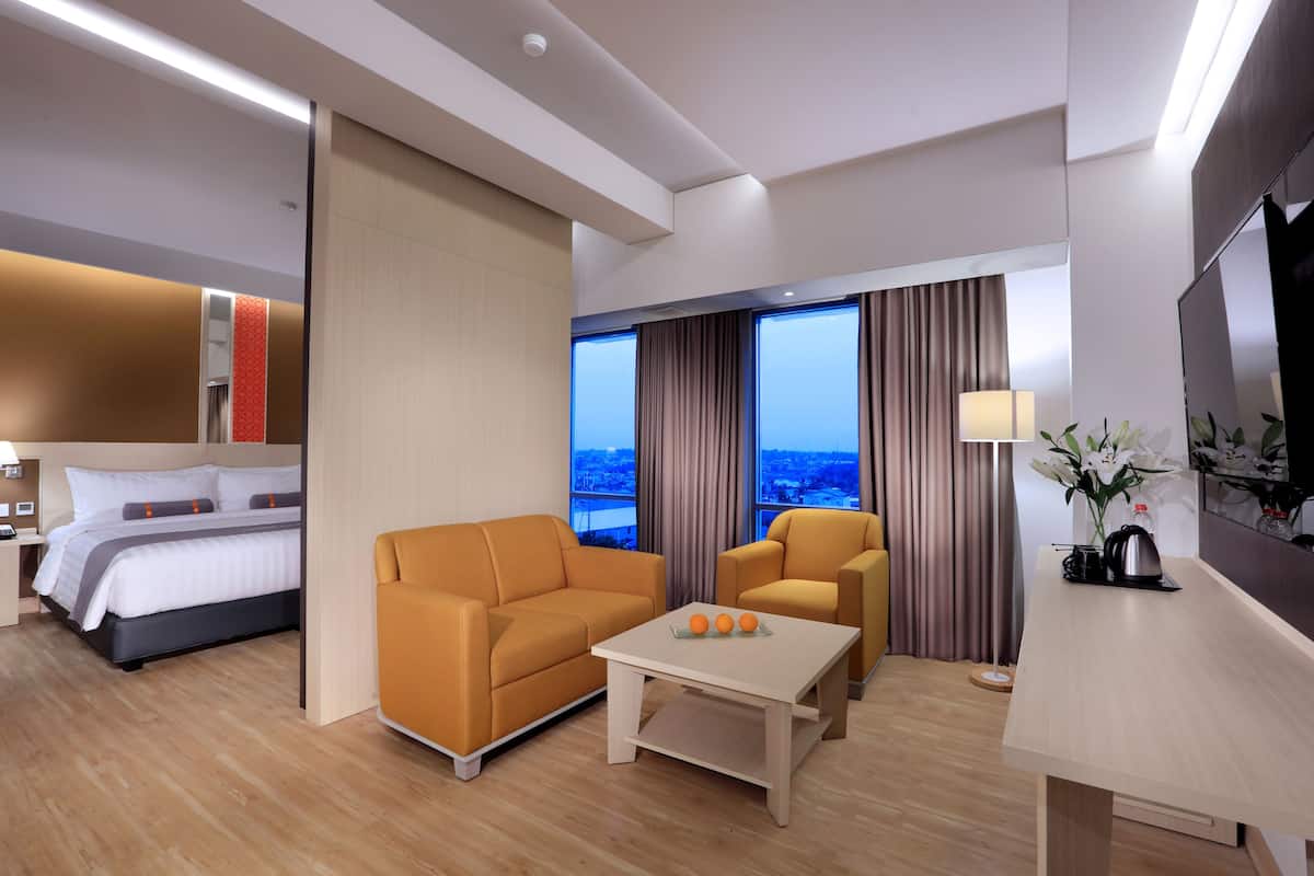 Quarto de hotel moderno com cama, dois sofás, mesa de centro, televisão e uma janela grande com vista para a cidade.
