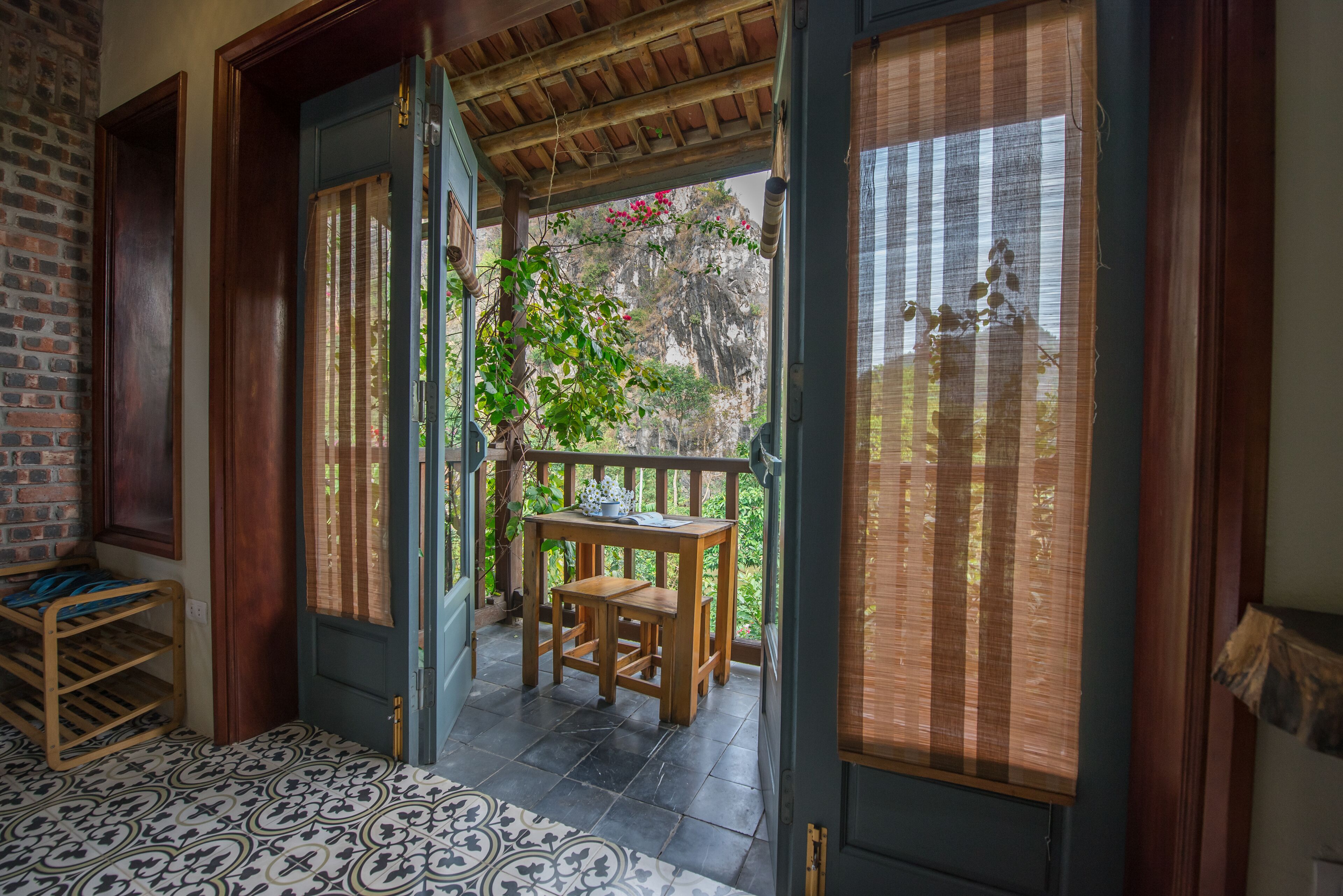 A wooden door leading to a balcony with a table and chairs, a patterned floor, and a view of greenery outside.