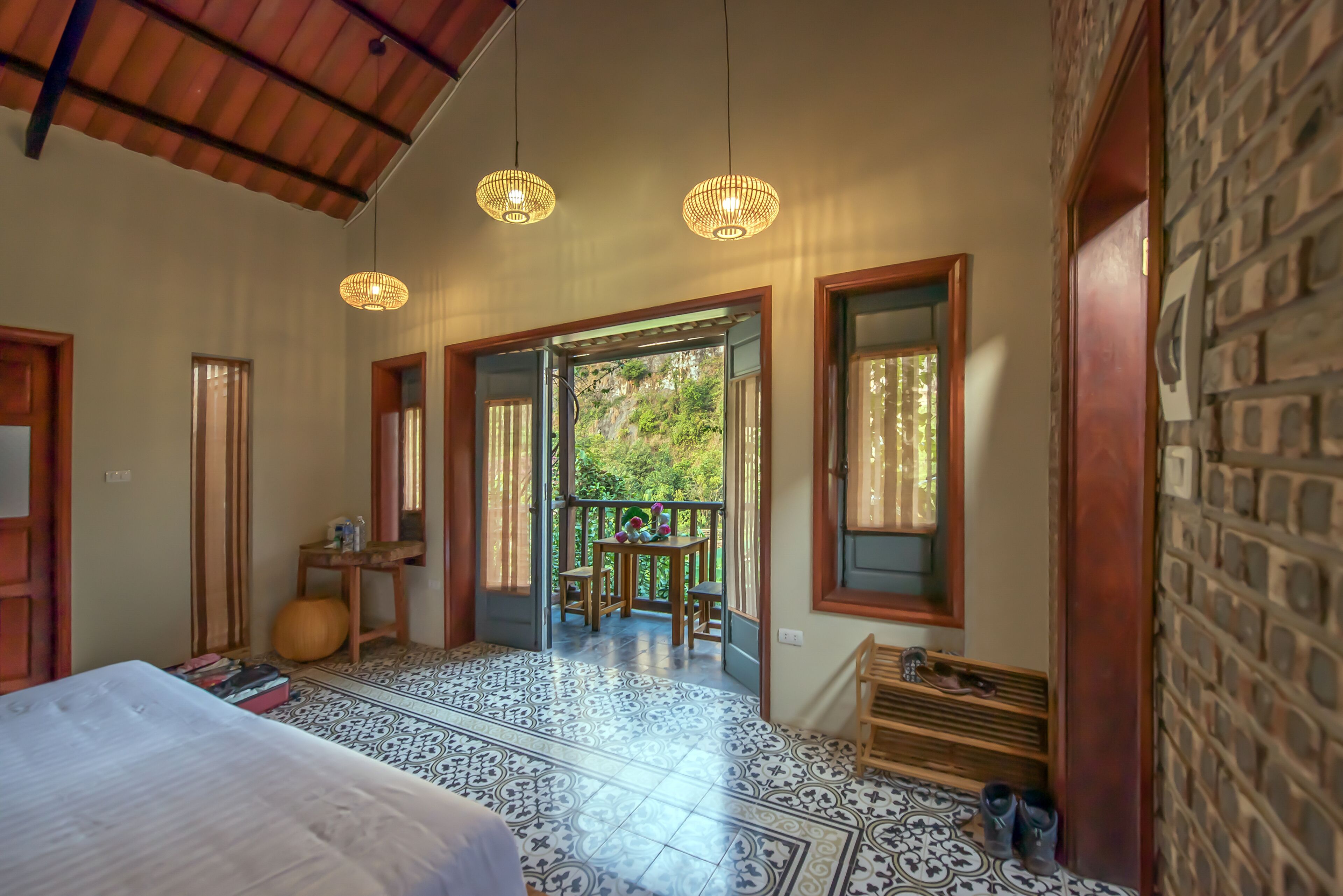 A room with a bed, a wooden bench, a table with chairs, and a balcony with a view of greenery.