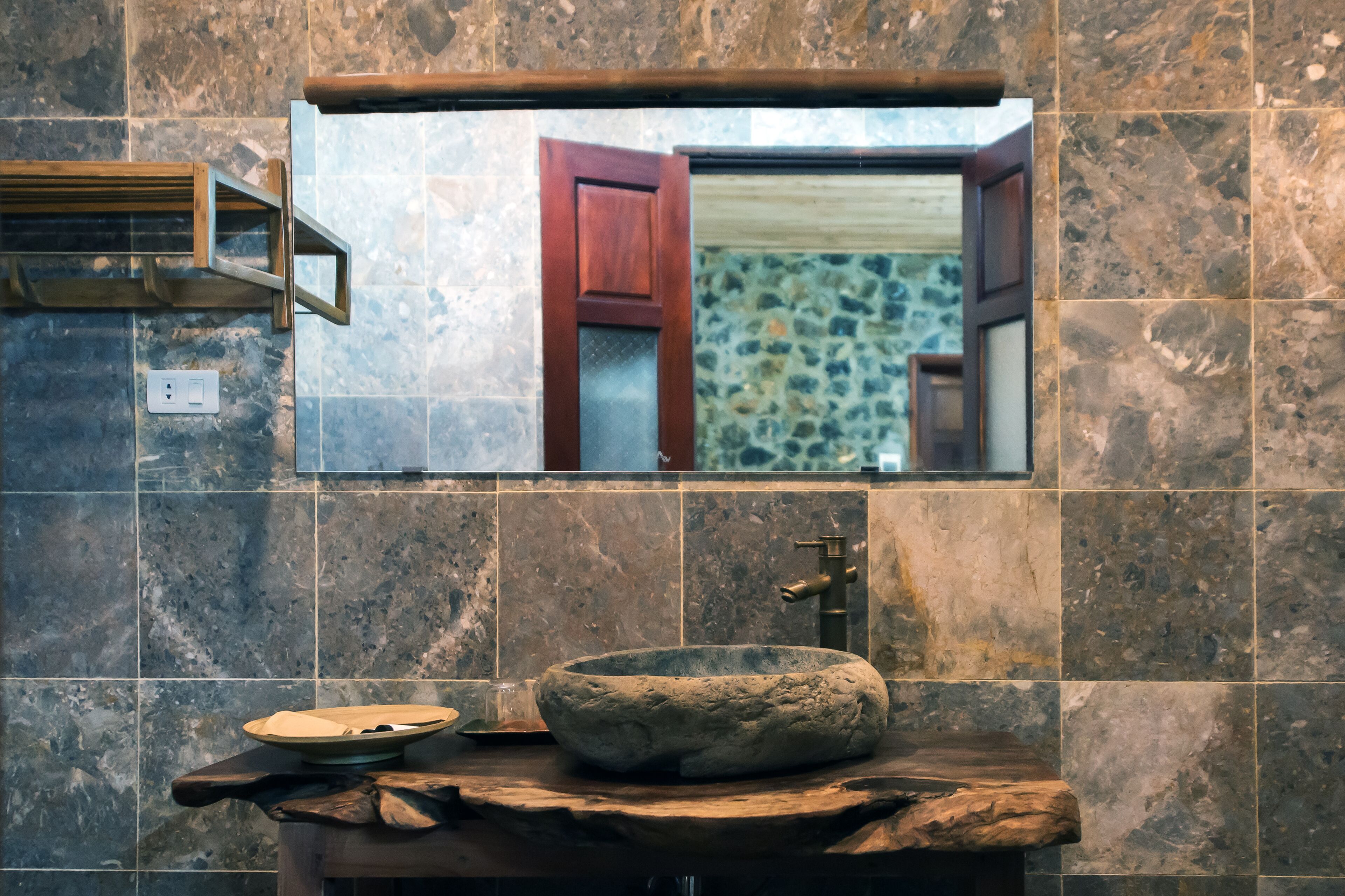 A bathroom with a stone sink, a wooden shelf, and a mirror reflecting a room with a door.