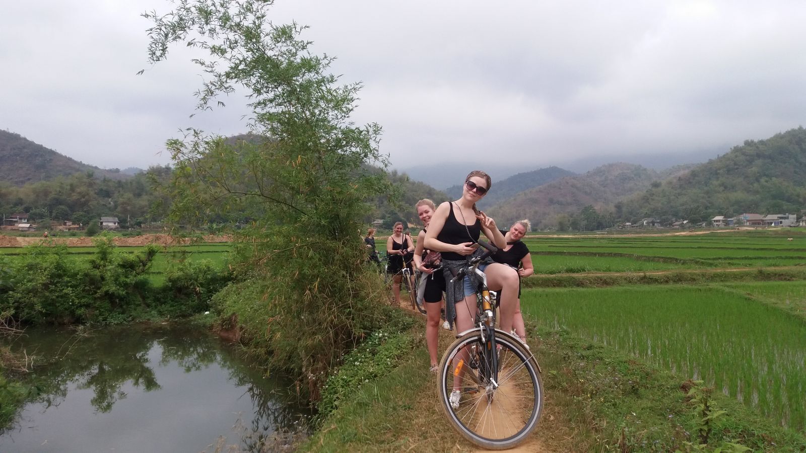 Un groupe de personnes fait du vélo le long d’une berge, avec des rizières et des montagnes en arrière-plan.