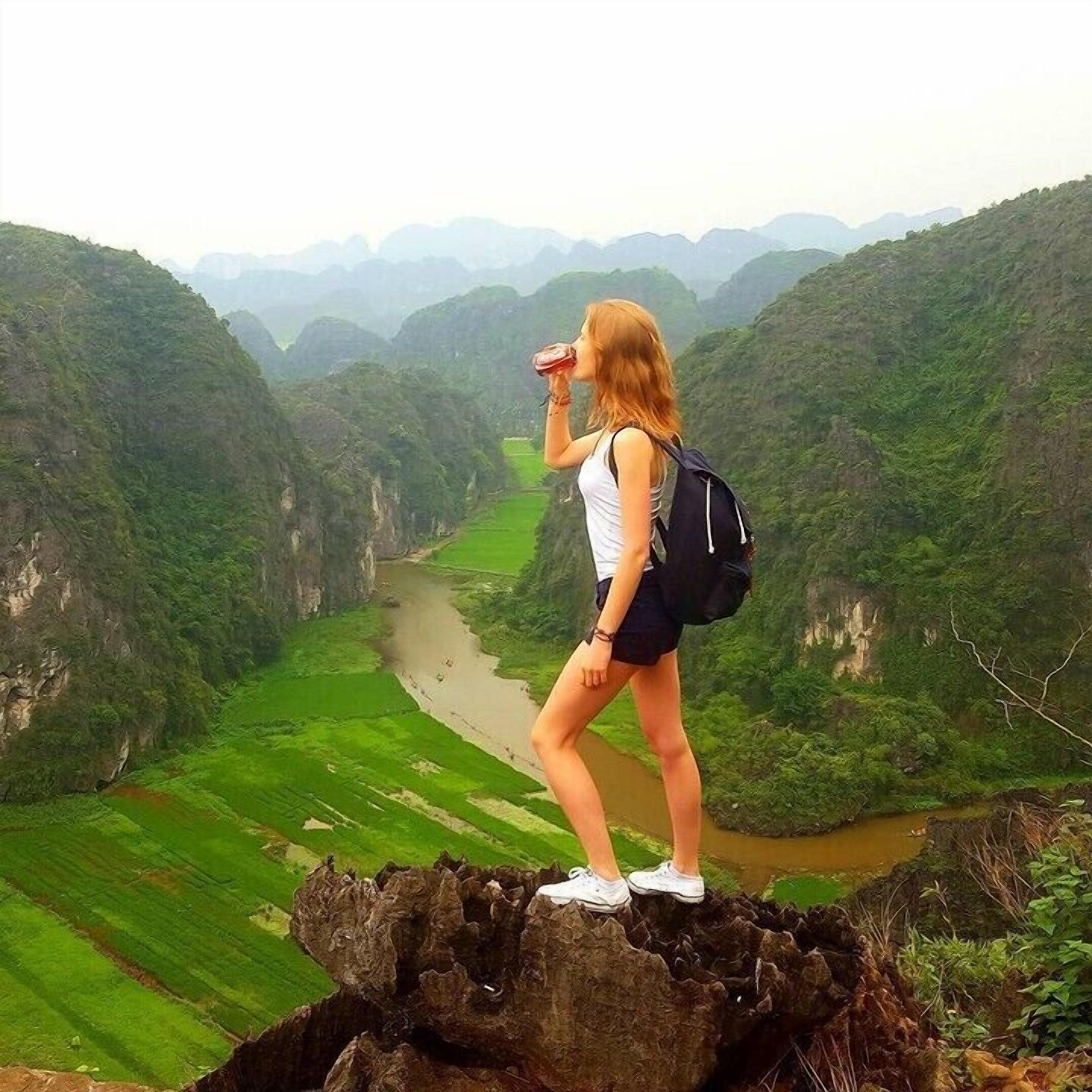 A person standing on a rocky outcrop overlooking a river valley with lush greenery and distant mountains.