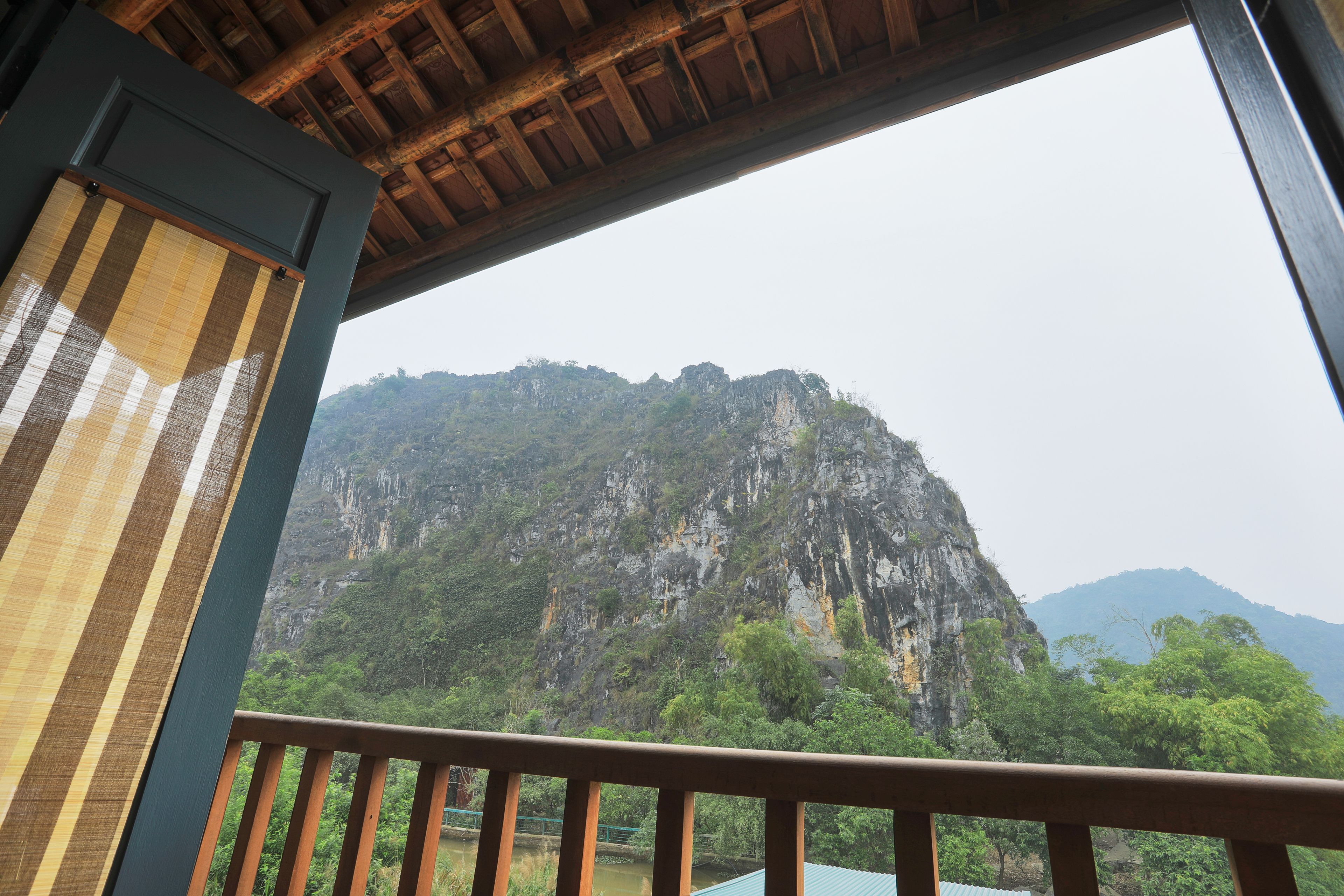 A view from a balcony with a wooden railing, overlooking a mountainous landscape with dense greenery and rocky cliffs.