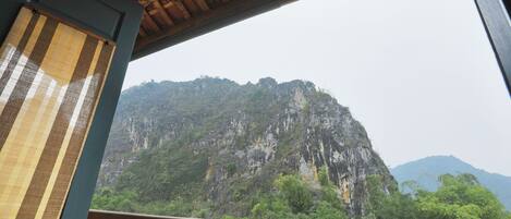 A view from a balcony with a wooden railing, overlooking a mountainous landscape with dense greenery and rocky cliffs.