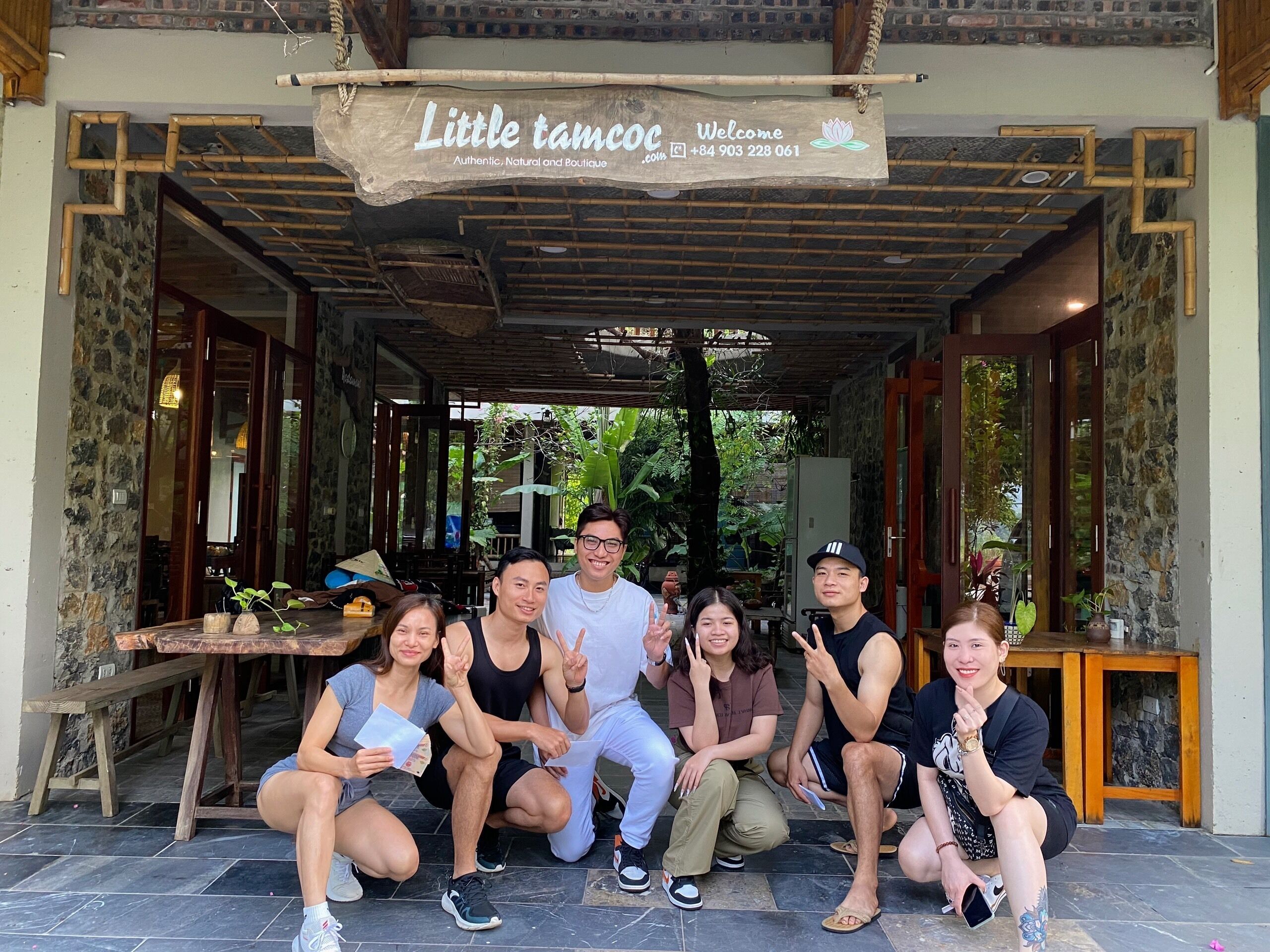 A group of people posing in front of a boutique hotel named "Little tamcoG" with a wooden signboard, stone walls, and a covered entrance.