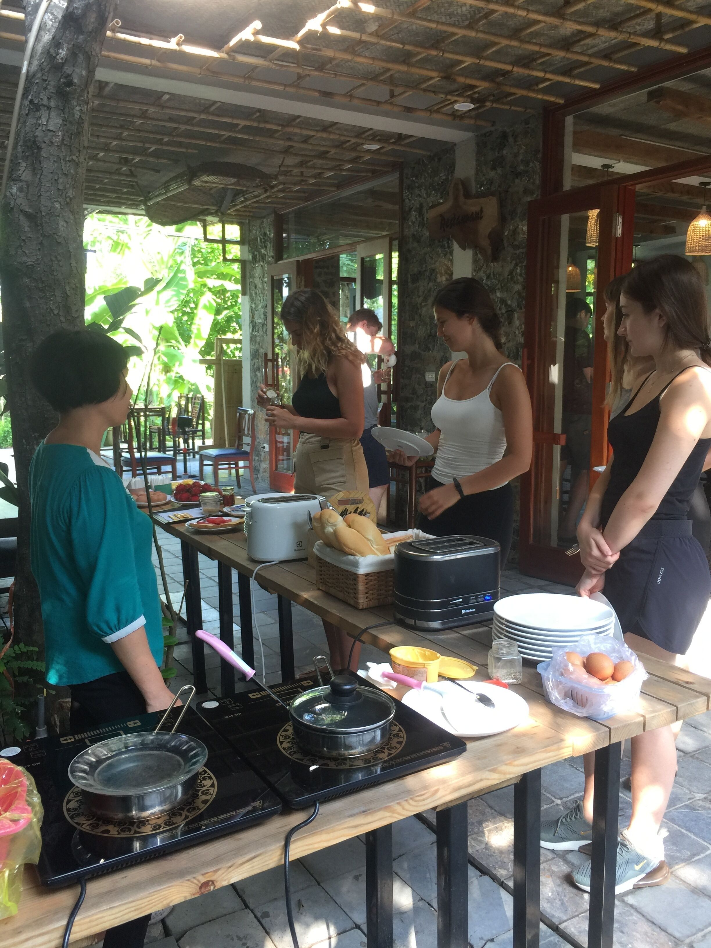 Un grupo de personas en un área de comedor al aire libre, con techo de bambú, pared de piedra y vista a la vegetación. Hay una mesa con comida, una estufa portátil y una cesta de pan.