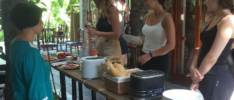 Un groupe de personnes est assis dans un espace de restauration en plein air, sous un plafond en bambou, avec un mur en pierre et une vue sur la verdure. On y voit une table avec de la nourriture, un réchaud portable et un panier de pain.