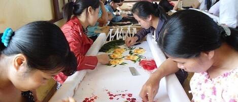 Un grupo de personas participan en una actividad artesanal, quizás bordando o pintando, en una habitación con vigas de madera en el techo y una ventana a la izquierda.