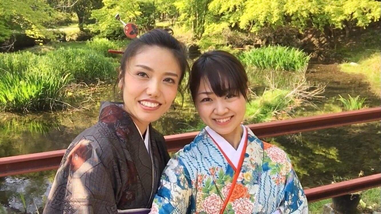 Two individuals in traditional attire standing on a bridge with a natural landscape in the background.