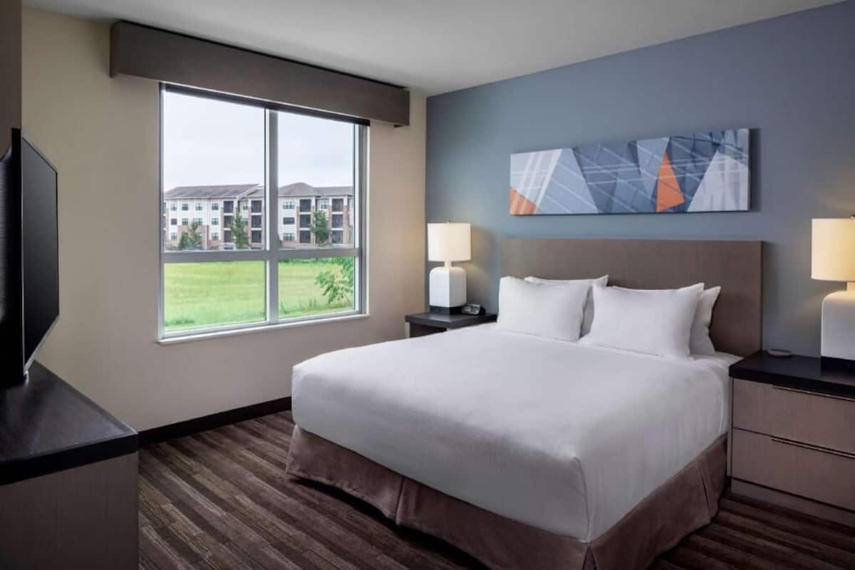 A hotel room with a large bed, two bedside tables, a TV, and a window with a view of buildings.