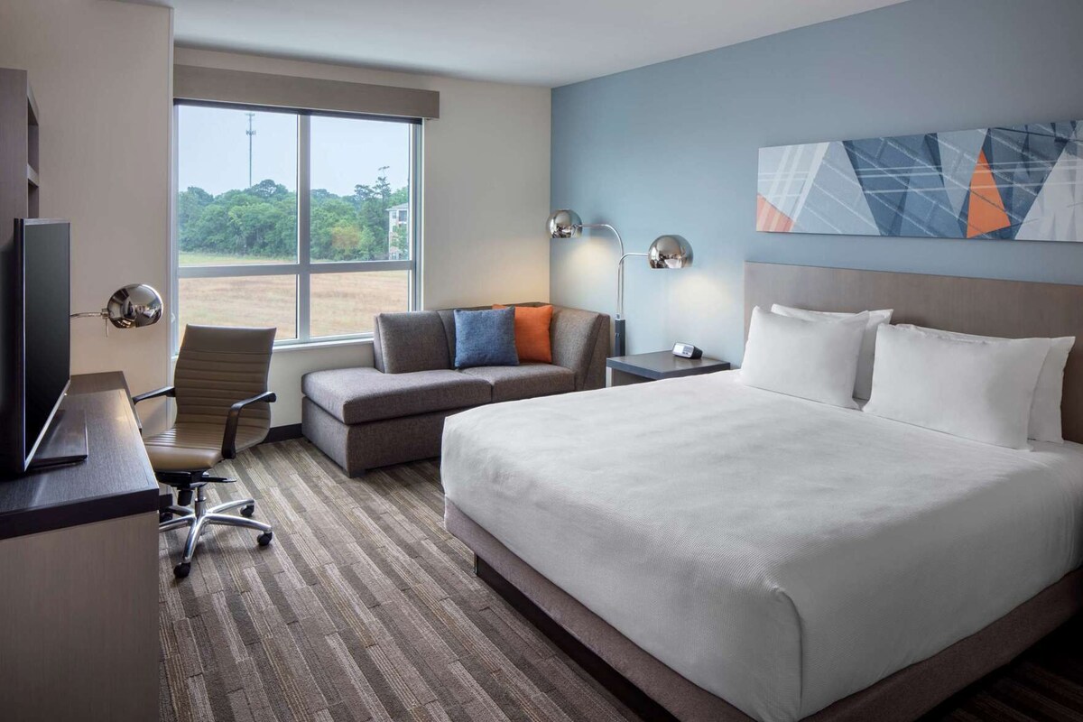 A hotel room with a large bed, a desk with a computer, a chair, a sofa, a TV, and a window with a view of trees and buildings.