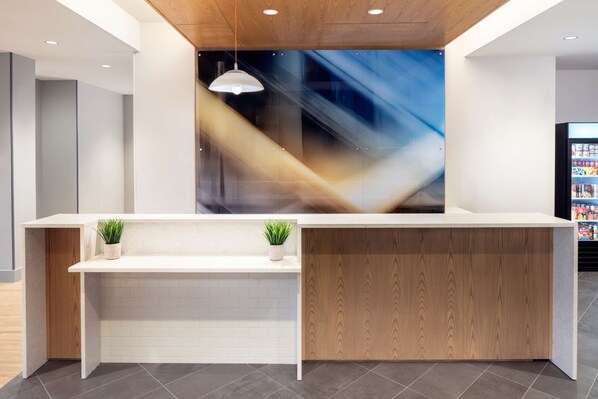 A reception desk with a modern design, featuring a white counter, two potted plants, and a wooden panel on the right. - Hyatt House Charleston / Mount Pleasant (Mount Pleasant)