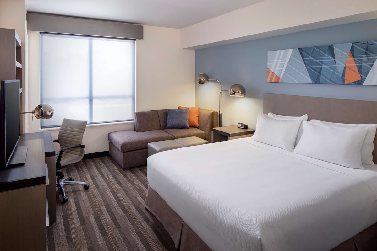 A hotel room with a large bed, a desk with a computer, a sofa, and a window with blinds.