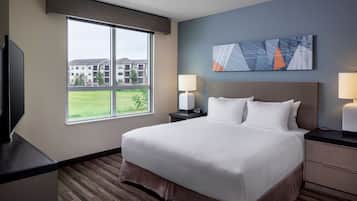 A hotel room with a large bed, two bedside tables with lamps, a flat-screen TV, and a window offering a view of the building and greenery outside.