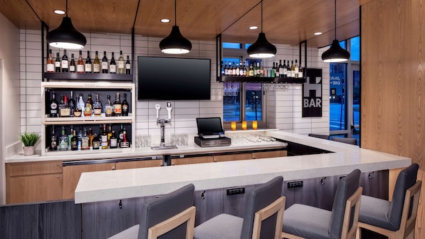 Eine moderne Bar mit einer langen Theke, mehreren Regalen voller Spirituosenflaschen, einem Flachbildfernseher und Barhocker.