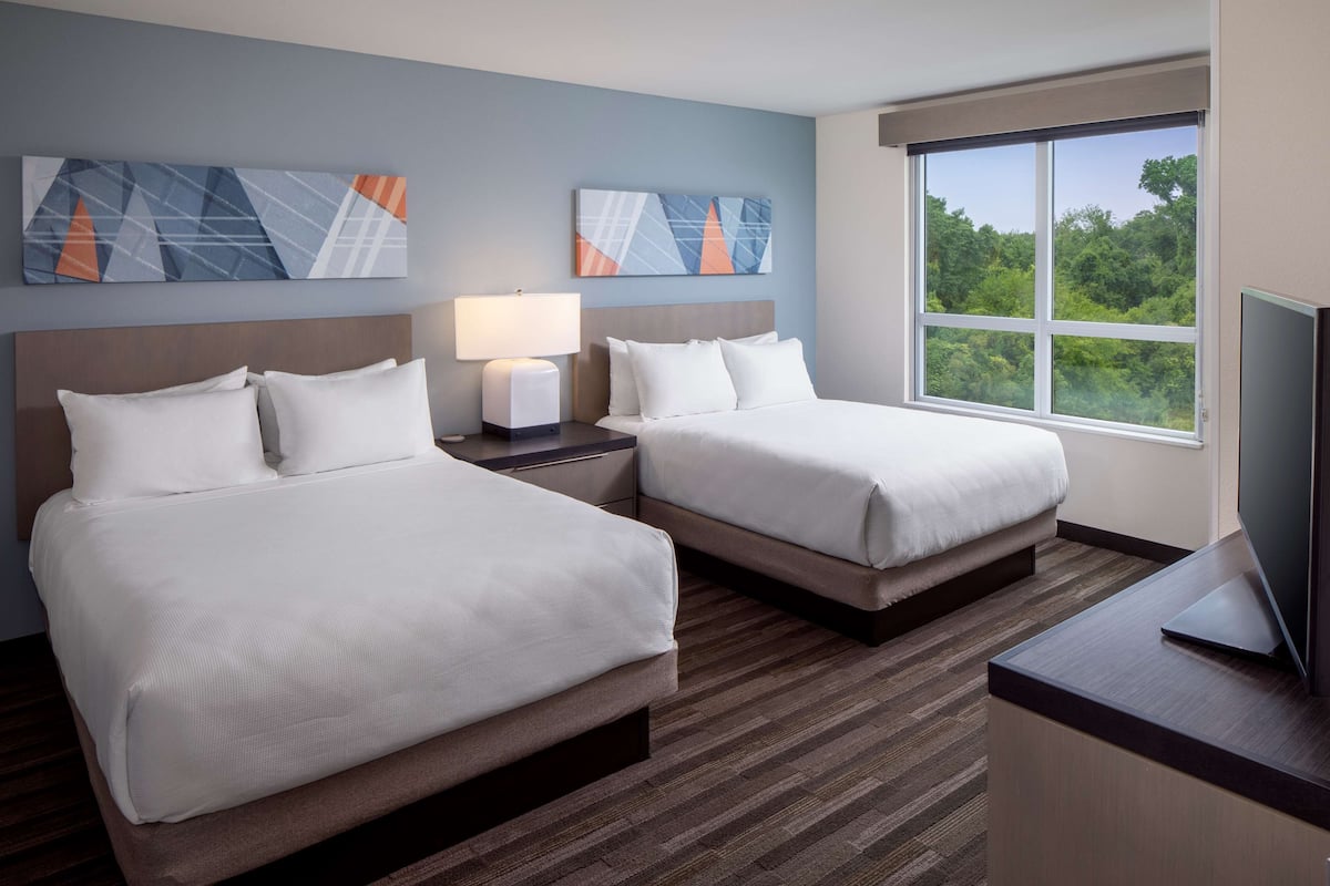 A hotel room with two beds, a nightstand with a lamp, a TV on a desk, and a large window with a view of trees.