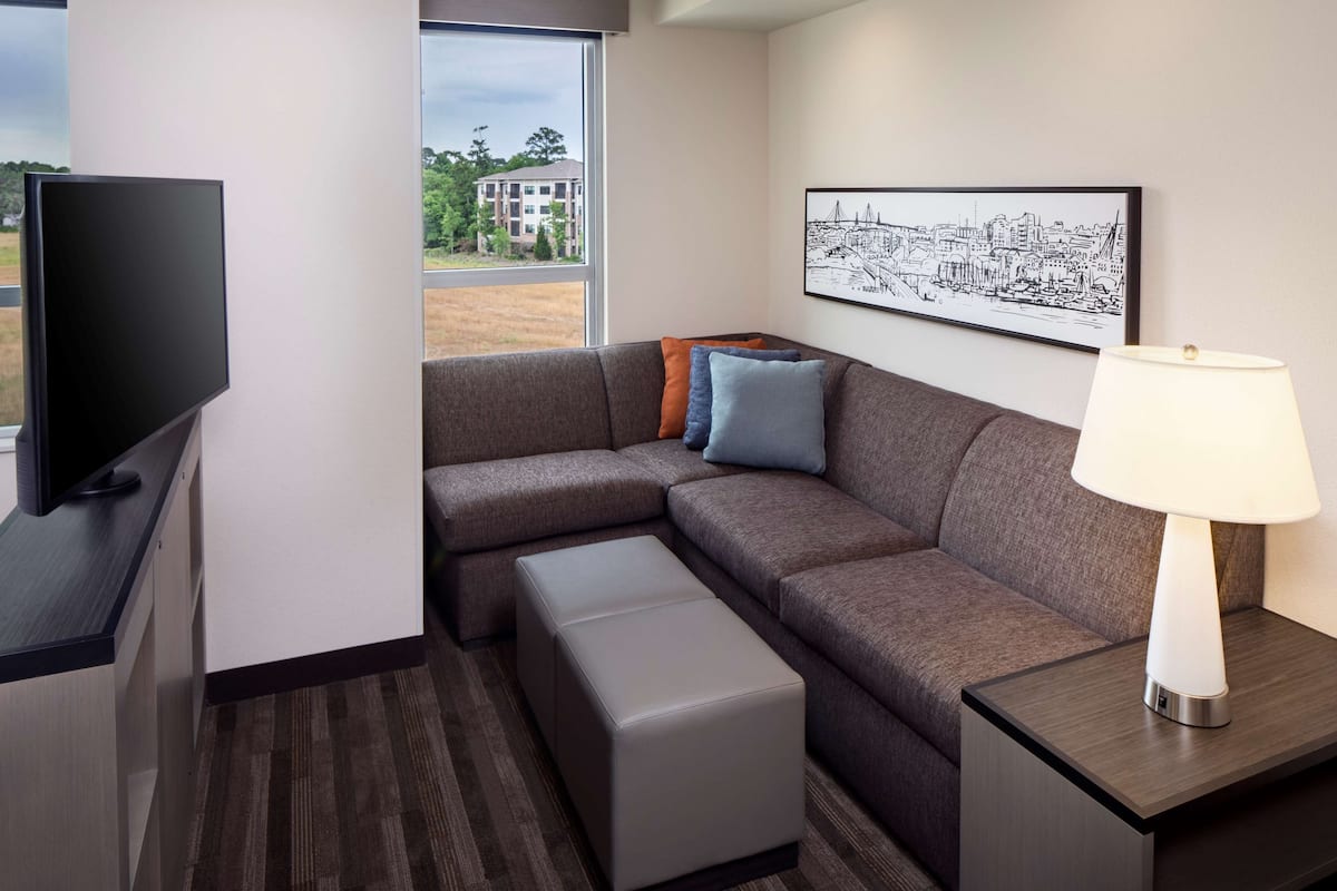 A modern hotel room with a sectional sofa, a flat-screen TV, a bedside table with a lamp, and a cityscape artwork on the wall.
