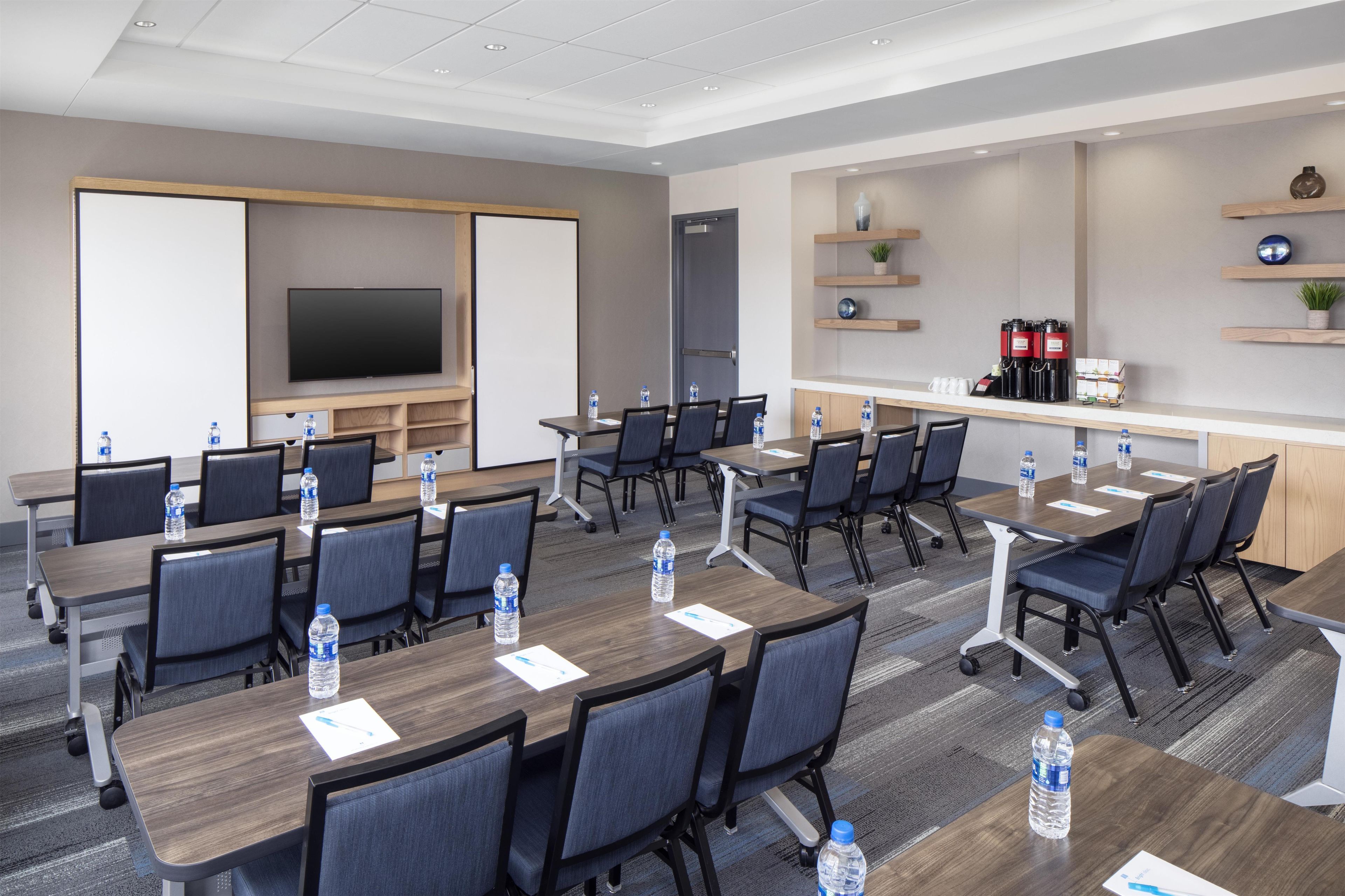 A conference room with multiple tables and chairs, equipped with water bottles, a flat-screen TV, and a coffee station.