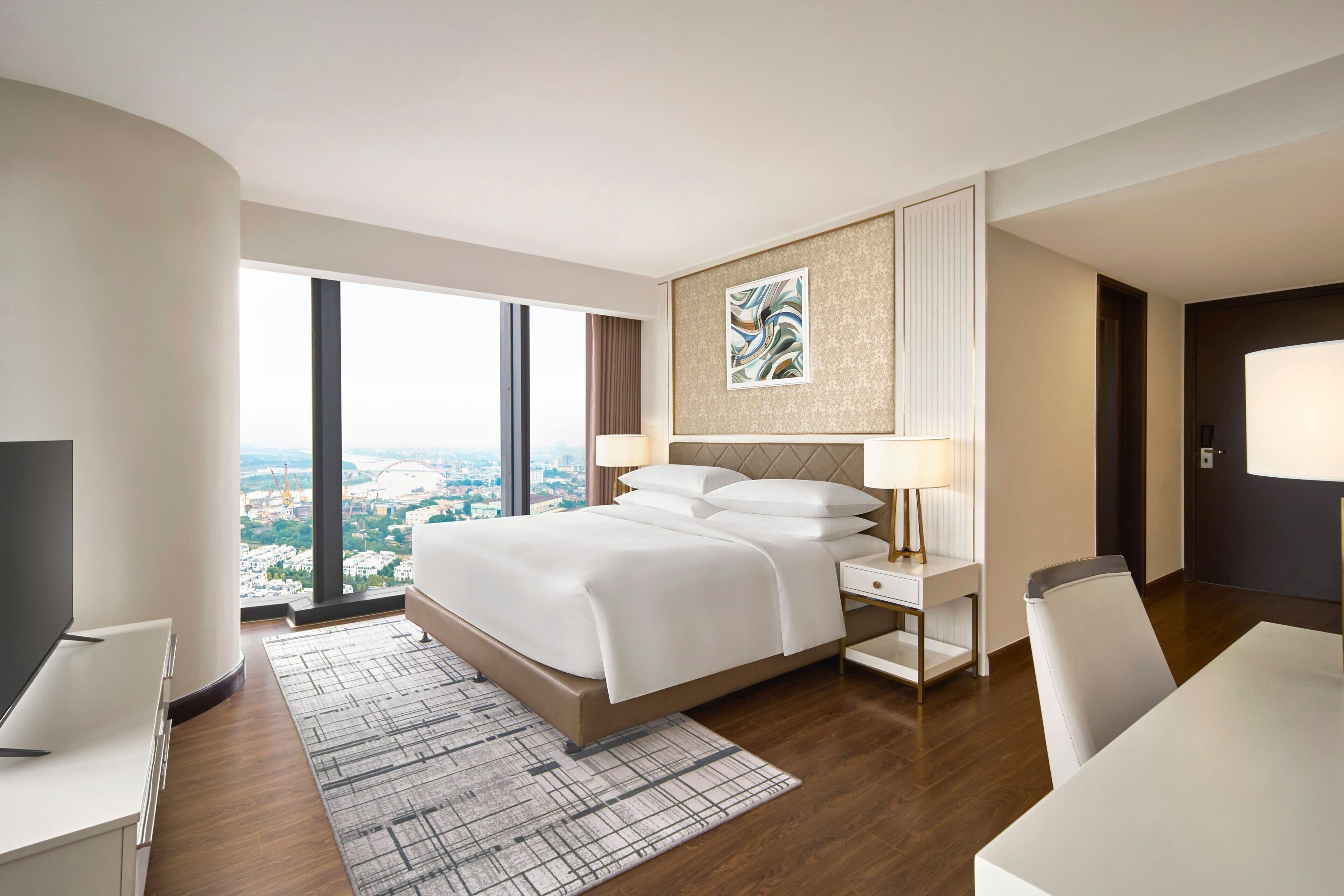 A modern hotel room with a large bed, a desk, a chair, a TV, and a view of the city through large windows.